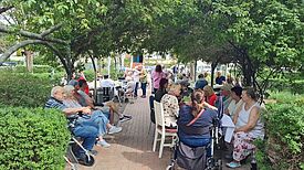 Mehrere ältere Menschen und Gäste sitzen und unterhalten sich bei einem Treffen unter schattigen Bäumen im Garten des Seniorenwohnhauses Voliére.