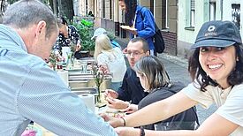 Mehrere Menschen sitzen und stehen an einer langen gedeckten Tafel im Freien und Essen gemeinsam.