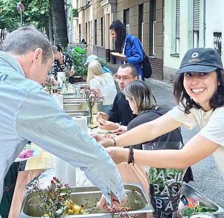 Mehrere Menschen sitzen und stehen an einer langen gedeckten Tafel im Freien und Essen gemeinsam.