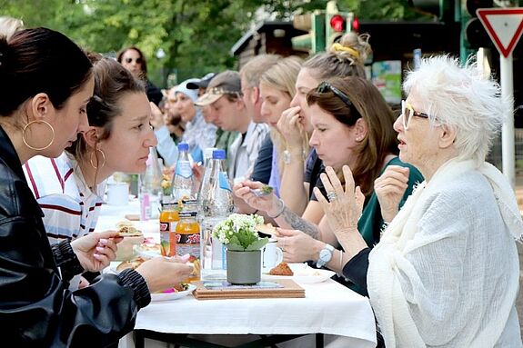 Zahlreiche Personen sitzen an einem langen und schmalen Tisch und unterhalten sich.