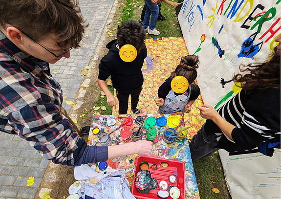 Kinder stehen vor einem Tisch mit Farbe und bemalen eine große Leinwand in Freien. 