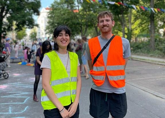 Kinder spielen auf einer für den Autoverkehr gesperrten Straße; Nachbarinnen und Nachbarn kommen zusammen, Straßenmöbel und Spielgeräte stehen im Bereich, Absperrungen und bunte Dekorationen sind sichtbar.