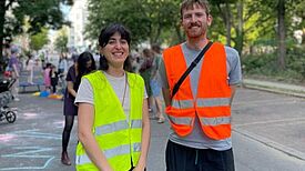 Kinder spielen auf einer für den Autoverkehr gesperrten Straße; Nachbarinnen und Nachbarn kommen zusammen, Straßenmöbel und Spielgeräte stehen im Bereich, Absperrungen und bunte Dekorationen sind sichtbar.