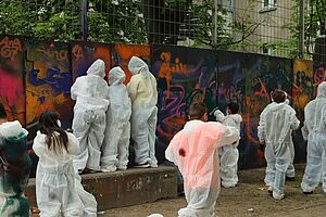 Viele junge Menschen vor einer bunt besprühten Wand. Sie sind in Rückenansicht erkennbar und tragen alle weiße Maleroveralls mit Farbklecksen. Manchen laufen, manche schauen auf die Wand und malen oder sprühen mit Farbe.