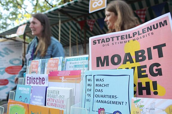 Broschüren und Flyer über das Quartiersmanagement Moabit-Ost liegen an einem Infostand aus, im Hintergrund stehen zwei Personen im Gespräch beim Perlenkiezfest 2025.