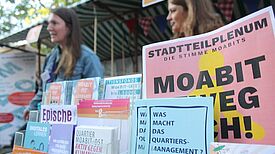 Broschüren und Flyer über das Quartiersmanagement Moabit-Ost liegen an einem Infostand aus, im Hintergrund stehen zwei Personen im Gespräch beim Perlenkiezfest 2025.