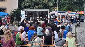 Menschen sitzen und stehen auf einem Platz vor einer kleinen Bühne mit Musikanlage und Schlagzeug. Im Hintergrund befinden sich Bäume und ein Banner, auf dem Fête de la Musique steht.