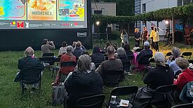 Menschen sitzen auf Stühlen vor einer Leinwand auf einer Wiese und schauen sich einen Film an. 