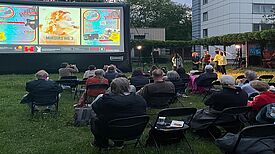 Menschen sitzen auf Stühlen vor einer Leinwand auf einer Wiese und schauen sich einen Film an. 
