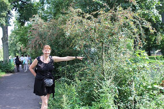 Eine Frau steht neben einer grünen Hecke und zeigt mit dem Finger auf diese.