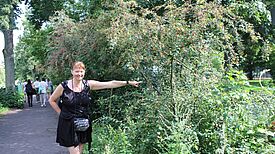 Eine Frau steht neben einer grünen Hecke und zeigt mit dem Finger auf diese.