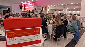 Eine rote Box mit der Aufschrift „Wie gut kennen Sie das Berliner Quartiersmanagement?“ steht im Vordergrund eines festlich beleuchteten Saals mit vielen Gästen an weißen Tischen.