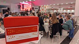 Eine rote Box mit der Aufschrift „Wie gut kennen Sie das Berliner Quartiersmanagement?“ steht im Vordergrund eines festlich beleuchteten Saals mit vielen Gästen an weißen Tischen.