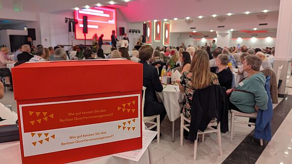 Eine rote Box mit der Aufschrift „Wie gut kennen Sie das Berliner Quartiersmanagement?“ steht im Vordergrund eines festlich beleuchteten Saals mit vielen Gästen an weißen Tischen.