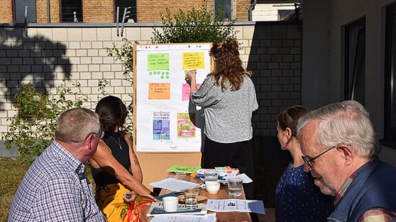 Mehrere Menschen sitzen bei sonnigem Wetter an einem Tisch im Freien, während eine Frau Notizzettel an eine Stellwand heftet.