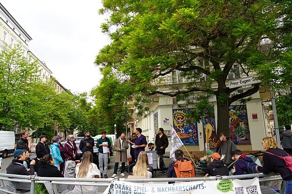 Eine Gruppe von Menschen versammelt sich beim Aktionstag zum Thema Klimaschutz unter einem großen Baum an der Ecke Prinz-Eugen-Straße, umgeben von bunten Wandbildern und Infomaterialien.