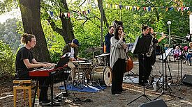 Eine Musikgruppe spielt im Freien mit Keyboard, Schlagzeug und Saxophon. Im Hintergrund sind Bäume zu sehen.