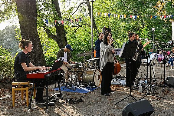 Eine Musikgruppe spielt im Freien mit Keyboard, Schlagzeug und Saxophon. Im Hintergrund sind Bäume zu sehen.