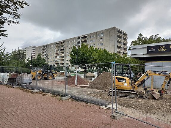 Eine eingezäunte Baustelle im Kosmosviertel mit Baggern und Radlader liegt vor einer Zeile von Wohnhäusern, Erdhaufen und Baustoffe sind gelagert.