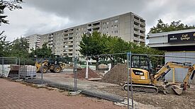 Eine eingezäunte Baustelle im Kosmosviertel mit Baggern und Radlader liegt vor einer Zeile von Wohnhäusern, Erdhaufen und Baustoffe sind gelagert.