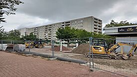 Eine eingezäunte Baustelle im Kosmosviertel mit Baggern und Radlader liegt vor einer Zeile von Wohnhäusern, Erdhaufen und Baustoffe sind gelagert.