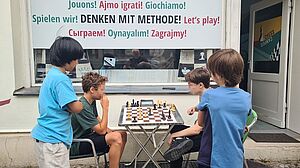 Vor einem Ladengeschäft spielen zwei Kinder Schach an einem Tisch. Vor jedem der Spielenden steht ein weiteres Kind und schaut dem Spiel zu. Am Fenster hängt ein Banner, auf dem in mehreren Sprachen steht: „Spielen wir! Denken mit Methode!“