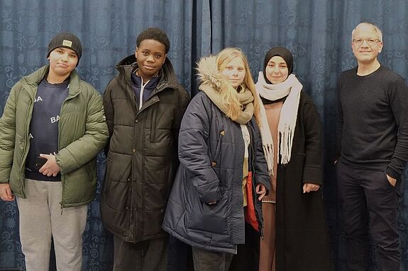 Vier Jugendliche und ein Erwachsener stehen vor einem blauen Vorhang.