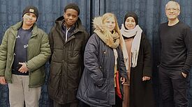 Vier Jugendliche und ein Erwachsener stehen vor einem blauen Vorhang.