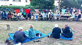 Menschen sitzen auf Picknickdecken und an langen Tischen im Freien unter bunten Wimpelketten.