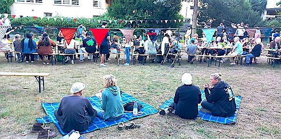 Menschen sitzen auf Picknickdecken und an langen Tischen im Freien unter bunten Wimpelketten.