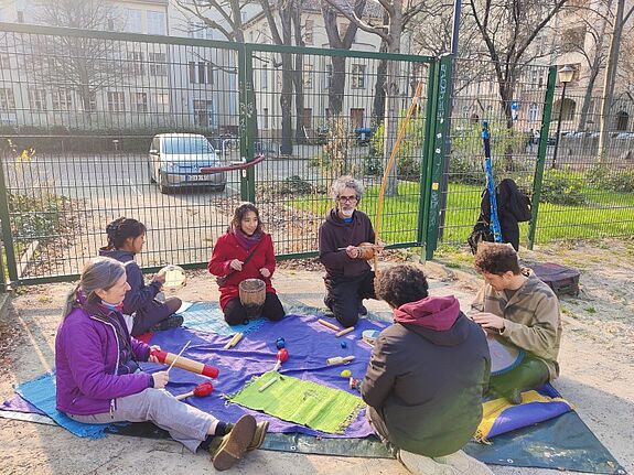Eine Gruppe von Menschen sitzt auf einer Decke im Freien und spielt mit Musikinstrumenten aus verschiedenen Kulturen.