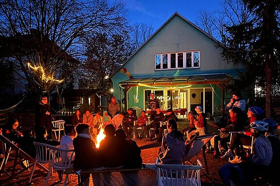 Kinder und Eltern sitzen am Lagerfeuer vor einem Haus und lauschen Märchen.