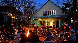 Kinder und Eltern sitzen am Lagerfeuer vor einem Haus und lauschen Märchen.