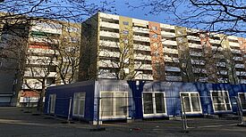 Auf dem Bild sieht man ein Hochhaus, vor dem das temporäre Stadtteilzentrum in einem blauen Containerbau untergebracht sein wird.  