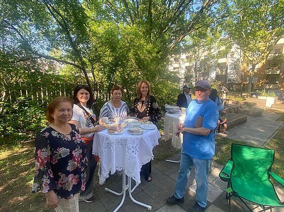Menschen unterschiedlichen Alters stehen bei sonnigem Wetter an Stehtischen mit gedecktem Kaffee- und Kuchenbuffet im Garten.