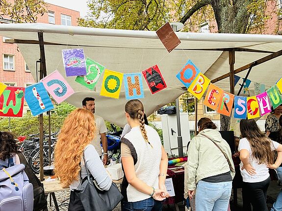 Mehrere Menschen stehen vor einem Stand mit weißer Dachplane und bunter Buchstaben-Wimpelkette. 