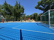 Im Vordergrund füllt ein leuchtend blauer Bolzplatz mit Toren und Fangzaun fast das ganze Bild. Dahinter stehen ein Spielbereich mit Klettergerüst und Rutsche sowie hohe Bäume.