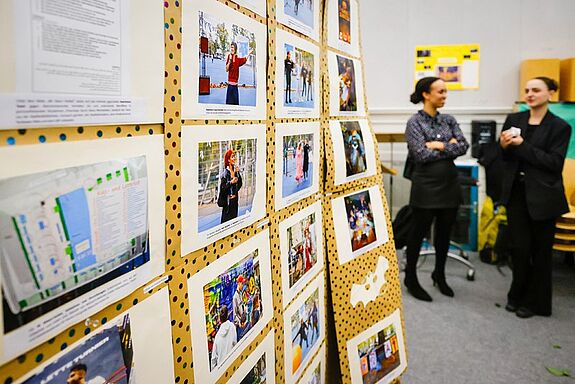 An einer Stelltafel hängen Bilder von Projekten aus dem QM, im Hintergrund sind zwei Personen zu sehen, die miteinander sprechen. 