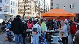 Menschen stehen vor einem orangefarbenen Zelt der Berliner Stadtreinigung und sortieren Gegenstände auf Tischen.