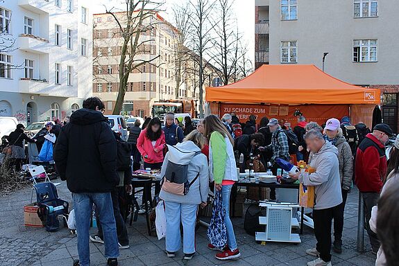 Menschen stehen vor einem orangefarbenen Zelt der Berliner Stadtreinigung und sortieren Gegenstände auf Tischen.