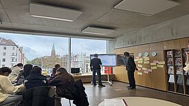 In einem Raum stehen zwei Personen vor einem Whiteboard und Post-its. Im Vordergrund sitzen mehrere Schülerinnen und Schüler.
