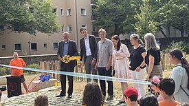 Sechs Erwachsene stehen auf einer frisch gepflasterten Fläche hinter einem blauen Band. Ein Mann hält einen großen gelben Schlüssel in der Hand. Im Vordergrund Kinder, im Hintergrund Bäume und ein Wohnhaus