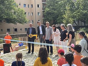 Sechs Erwachsene stehen auf einer frisch gepflasterten Fläche hinter einem blauen Band. Ein Mann hält einen großen gelben Schlüssel in der Hand. Im Vordergrund Kinder, im Hintergrund Bäume und ein Wohnhaus