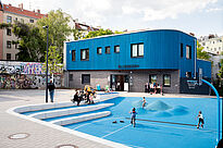 Im Hintergrund steht das Blueberry-Gebäude mit blauer Fassade. Davor spielen mehrere Kinder auf einem blauen befestigten Vorplatz mit wellenförmigen Bodenelementen, während Erwachsene und Kinder auf hellen Sitzstufen und vor dem Eingang sitzen.