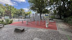 Parkour-Geräte stehen auf einer roten Fläche neben einem eingezäunten Ballspielplatz und Bäumen.