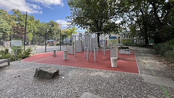 Parkour-Geräte stehen auf einer roten Fläche neben einem eingezäunten Ballspielplatz und Bäumen.