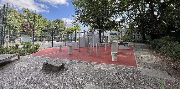 Parkour-Geräte stehen auf einer roten Fläche neben einem eingezäunten Ballspielplatz und Bäumen.