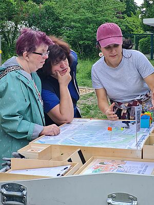 Drei Menschen beratschlagen sich über einer Karte und platzieren darauf verschiedenfarbige Fähnchen. Im Hintergrund grüne Umgebung.