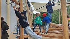 Mehrere Kinder hangeln sich entlang der Spielgeräte und balancieren auf Baumstämmen. 