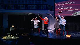 Vier Personen treten auf einer Bühne beim Neujahresempfangs des QMs auf, während sie eine Person auf dem Klavier begleitet.